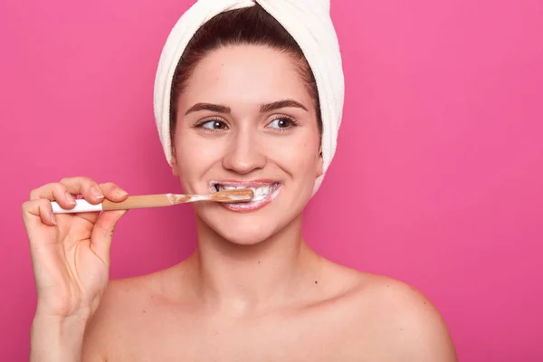 Retrato de una atractiva mujer caucásica sonriente cepillándose los dientes aislada sobre la pared rosa del estudio, de pie con una toalla blanca en la cabeza y hombros anudados, mirando a la distancia, sostiene el cepillo de dientes . — Foto de Stock