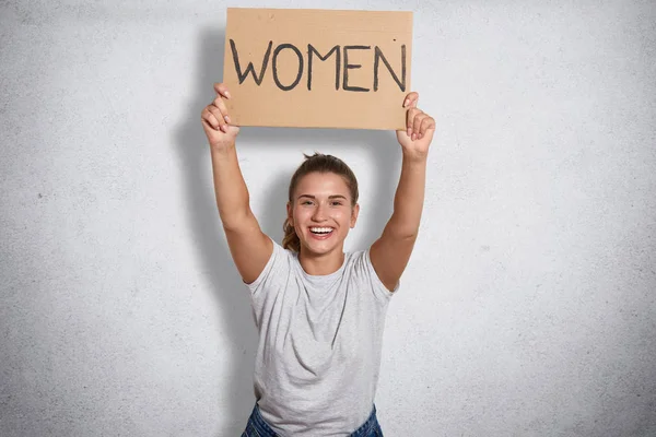 Cheerful good looking feminist smiling sincerely, raising inscription women in both hands, ready to public activity, peaceful demonstration, being in high spirits, wearing casual white clothes. — Stock Photo, Image