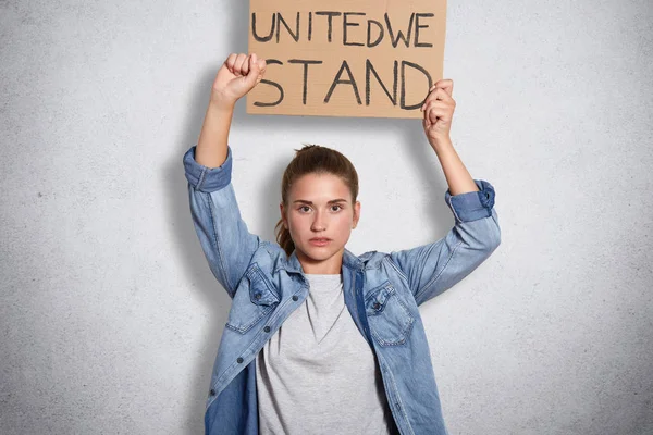 Portrait of militant powerful young feminist showing her fist, holding sign with inscription united we stand in one hand, having decisive facial expression, interested in women social problems.