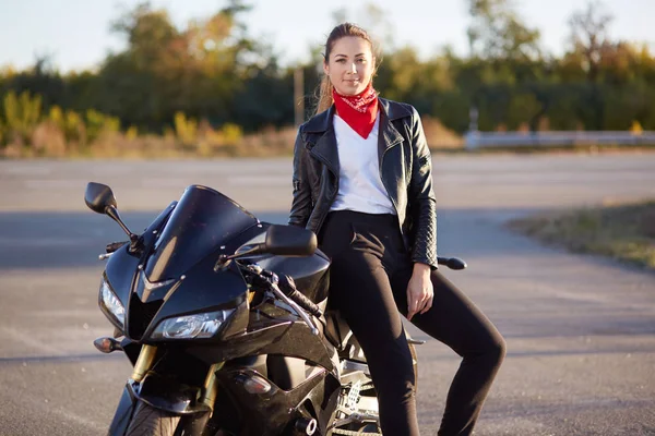 Tiro ao ar livre de mulher com cabelo escuro sentado em motobike preto, usa jaqueta de couro, calças, camisa branca e bandana vermelha, olhando sorrindo diretamente para a câmera, jovem gosta de alta velocidade . — Fotografia de Stock