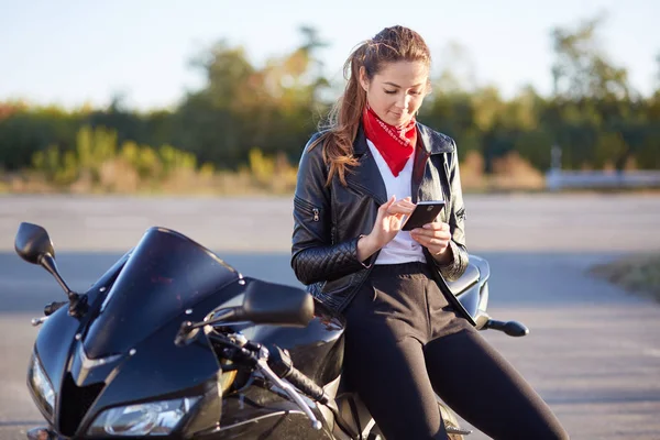 Young female biker with phone in hands, typing message on cellular, wears red bandana, white shirt and leather jacket, poses on motorbike. Technology and lifestyle, high speed, traveling on motorcycle — Stock Photo, Image