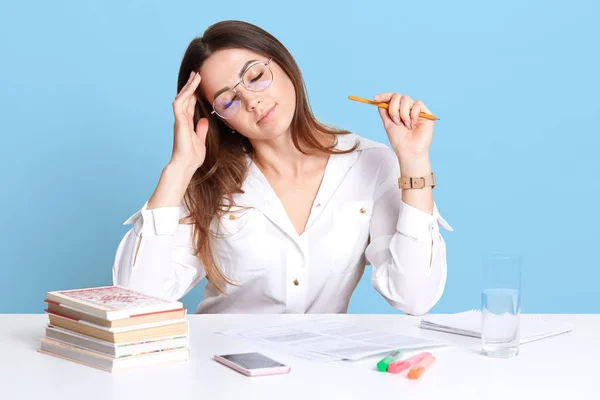 Indoor shot of exhausted young dark haired businesswoman sitting in office at white desk, keeps eyes closed, touching her forehead, looks sick and tired of working with papers. People and job concept. — Stock Photo, Image