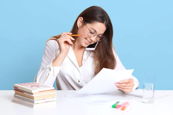 Studio intérieur tourné d'une élégante jeune femme assise au bureau blanc, parlant via smartphone tout en travaillant à son travail de cours, vêtue d'une blouse formelle, semble heureuse, isolée sur fond bleu . — Photo