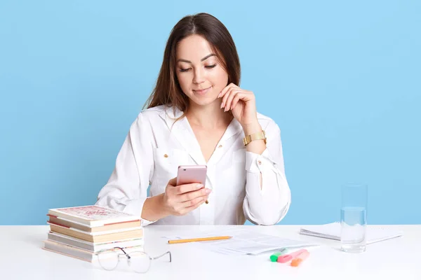 Indoor shot of young businesswoman sitting in office, touching her chin with fingers, holding smart phone in other hand, checking social net work, posing solated over blue background in studio. — Stock Photo, Image