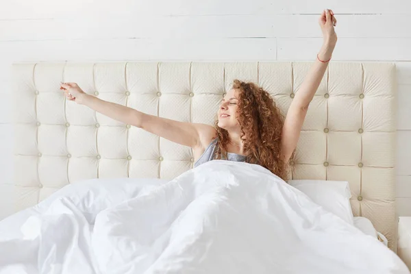 Adorable magnetic curly haired female sitting in her bed, covered by blanket, looking aside, being in high spirits, stretching, raising her arms up, smiling sincerely, getting up in morning. — Stock Photo, Image
