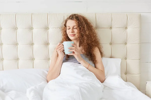 Photo of charming young woman with curly hair, being in bed, drinking aromatic coffee ii morning, look calm and happy, keeps eyes closed, relaxing in bedroom. Weekend, relax and harmony concept. — Stock Photo, Image