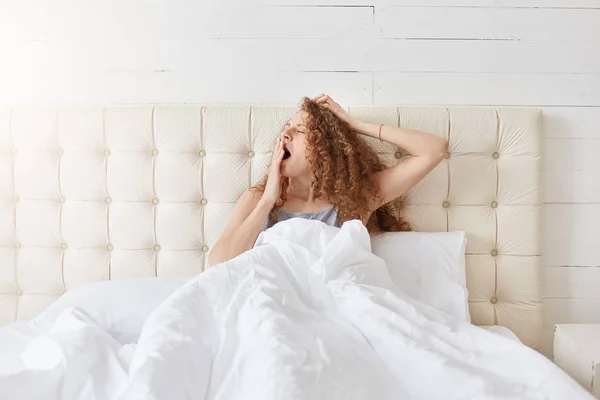 Indoor shot of peaceful attractive young lady relaxing in her bed in morning, yawning, covering her mouth with one hand, having curly hair, enjoying start of new day. People and morning concept. — Stock Photo, Image