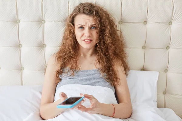 Horizontal shot of young female with incomprehensible emotions, girl with curly hair dressed in nightgown, lying in bed, looking directly at camera, using her smart phone for cheching social networks. — Stock Photo, Image