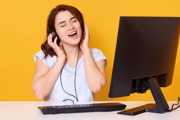 Studio shot of girl listening music with headphone and singing songs loudly, sitting at white desk in front of personal computer, lady dressed casual white t shirt, posing against yellow studio wall . — Stok Foto