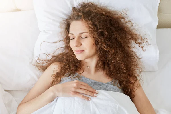 Indoor shot of young female with curly hair, laying in white bed with closed eyes and happy facial expression. Attractive woman stays in bed all day alone, has weekend. Happyness and rest concept. — Stock Photo, Image