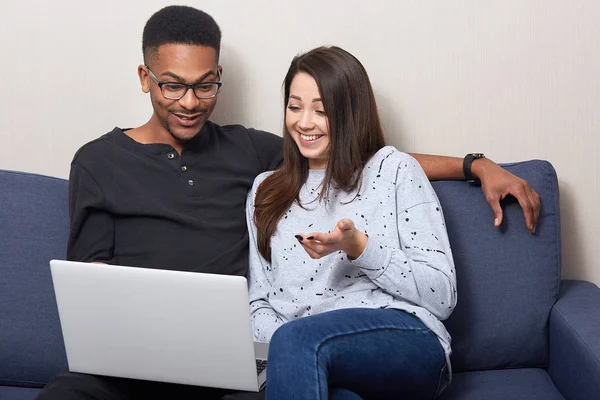 Smiling black young man and dark haired woman relax on couch, watching movie on laptop, happy multicultural couple resting on sofa during weekend, enjoying to spend time together. Relationship concept — Stock Photo, Image