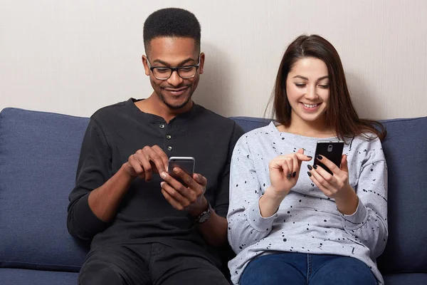 Photo of young woman in casual outfit sitting on couch in room with her African boyfriend and looking at theit smartphones's on smartphone screens and loughing, see something interesting, check email. — Stock Photo, Image