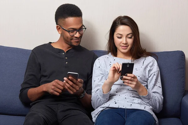 Multicultural couple sitting on sofa at home looking in theirs smart phones, using internet, being happy spending time together, woman showing something in cell phone. People and family time concept. — Stock Photo, Image