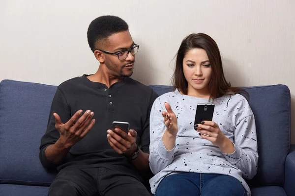 Studio shot of couple sitting together on comfortable sofa in apartment and using mobile phone, looking on screen, dark skinned male and caucasian female wearing casually, reading something unpleasant — Stock Photo, Image