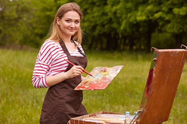 Retrato de menina alegre sorridente com longos cabelos loiros segurando seu pincel e paleta suja, criando obra-prima com inspiração, olhando diretamente para a câmera, estando em bom humor. Conceito de pintura . — Fotografia de Stock