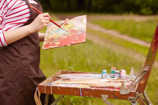 Tiro ao ar livre de mulher desconhecida em torno da natureza, vestindo avental marrom, segurando escova e paleta, fazendo pincelada varrendo, criando esboços ao ar livre, tubos com tintas estão em vista. Conceito de arte . — Fotografia de Stock