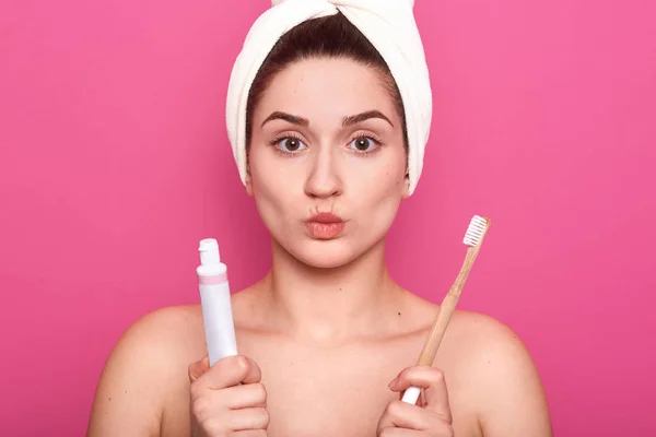 Studio shot van tevreden vrouwelijke model pouting lippen, draagt witte handdoek op het hoofd, klaar voor het poetsen van tanden met tandenborstel en tandpasta, zorgt voor hygiëne en schoonheid, geïsoleerd over roze achtergrond. — Stockfoto