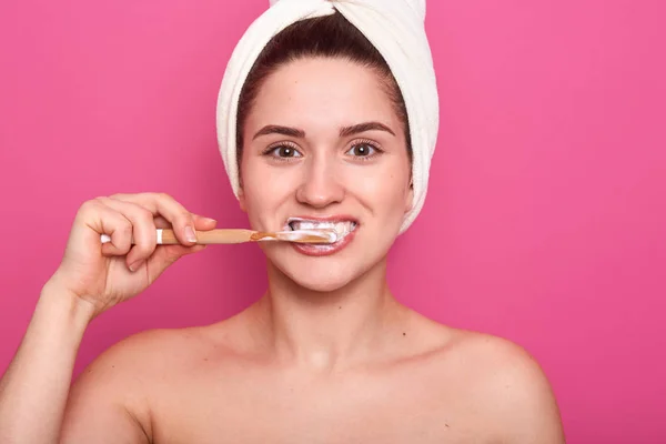 Vrouw met tandenborstel, schoonmaken van haar tanden, poseren in de badkamer met blote hoofd en witte handdoek, vrouw doet 's ochtends procedures na het nemen van een douche, geïsoleerd op een rooskleurige achtergrond. Hygiënisch concept. — Stockfoto