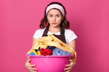 Hafif saç bandı, beyaz t shirt ve kahverengi önlük ile hayal kırıklığına duygusal kadın, çamaşır tutan, ev işleri yorgun, doğrudan kameraya bakarak. Ev hanımı ve çalışma konsepti.