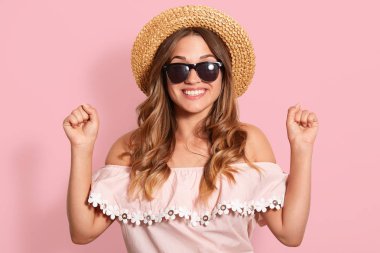 Yakın portre al kıyafet, hasır şapka ve siyah güneş gözlüğü, pembe arka plan üzerinde yumruklar içine ellerini sıkma, dişli gülümseme ile poz, mutlu yüz ifadesi vardır, onu succees kutluyor.