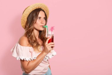 Genç güzel kadın Studio shjt, çıplak omuzve hasır şapka ile gömlek poz, pembe arka plan üzerinde izole tropik kokteyl içme. Reklam veya promosyon metniniz için alan kopyalama.