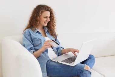 Neşeli iyi görünümlü model evde kanepeüzerinde poz, faaliyetleri aracılığıyla dizüstü bilgisayar ile meşgul olmak, online hizmetler çalışıyor, içtenlikle gülümseyerek, kredi kartı tutarak, satın almak için bir şey arıyor.