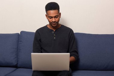 Evde online çalışan koyu tenli genç erkek Kapalı çekim, taşınabilir dizüstü bilgisayarda internet sörf, rahat mavi kanepede oturan, yakışıklı adam rahat giyinmiş. İnsanlar ve iş kavramı.