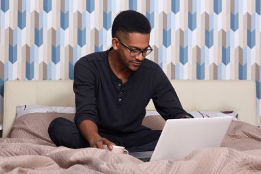 Görme düzeltme ve siyah rahat kıyafet için gözlük giyen koyu tenli adam, evde online çalışma, dizüstü bilgisayar ve kablosuz internet kullanarak, uzak iş zevk konsantre görünüyor.