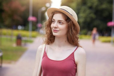 Yeşil parkta poz veren güzel ihale modelinin dış görüntüsü, yeşil alanda zaman geçirmek, doğadan etkilenmek, bir kenara bakmak, hasır şapka ve kırmızı üst takmak, yerel sokağın ortasında durmak.