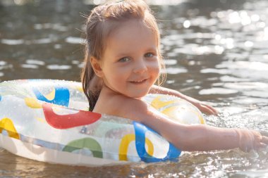 Samimi bir şekilde gülümseyen, hoş bir yüz ifadesine sahip, doğrudan kameraya bakan, nehirde yüzme, yüzme çemberine tutunma, yüksek ruhlu olmak gibi neşeli neşeli bir çocuğun portresini kapatın.