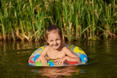 Özel ekipmanlarla nehirde yüzen, yüzmeyi öğrenecek, hoş bir yüz ifadesine sahip, içtenlikle gülümseyen, arka planda sazlık olan tatlı oyuncu çocuk portresi. Doğa kavramı.
