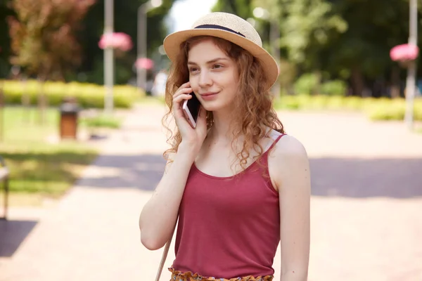 Plan horizontal de belle jeune femme en chapeau de paille et t-shirt bordeaux, être dans le parc de la ville, se reposer avec des amis, appeler son petit ami via smartphone, a expression faciale agréable . — Photo