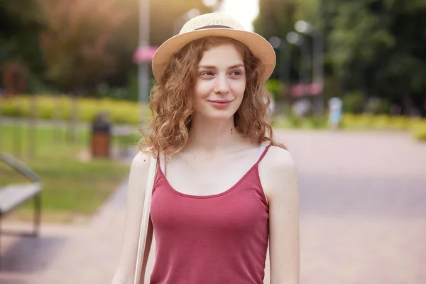 Fille romantique portant chapeau de paille et t-shirt bordeaux posant dans le parc, regardant souriant de côté, enjoignant belle nature. Adorable femme aux cheveux ondulés marche dans le parc, attendant son amie, ayant des vacances . — Photo