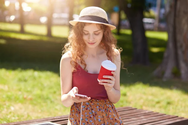 Image of woman with foxy hair sitting on bench in city park, charging mobile phone on bench via solar panel, holding takeaway coffee and checking her social network. Modern technology concept.