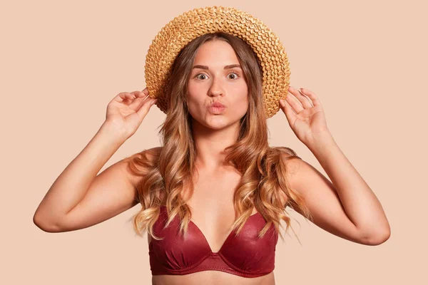 Close up portrait of attractive woman wearing red swimsuit on beige background and straw hat on her head, keeps hands on hat and posing with lips rounded, makes kiss gesture. Body language concept. — Stock Photo, Image