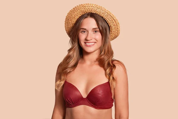 Primer plano retrato de mujer joven en ropa de playa roja y sombrero de paja, se encuentra sonriendo y mirando directamente a la cámara, posando aislado sobre fondo de estudio beige. Copiar espacio para la publicidad . — Foto de Stock
