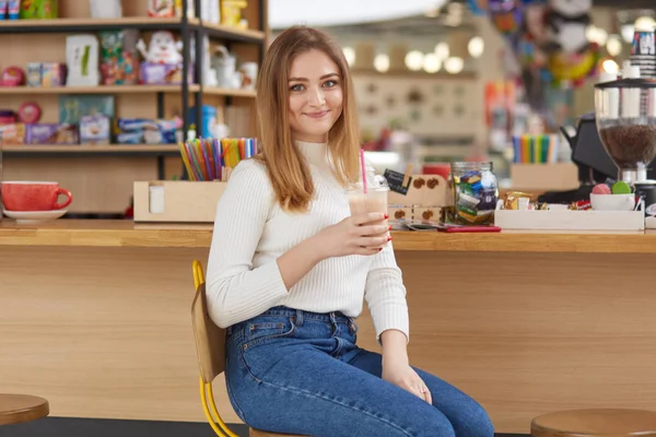 Indoor shot di giovane bella donna bionda che indossa camicia bianca casual e jeans, seduto al bar o al caffè, bere cocktail, studente femmina ha pausa tra le lezioni, ha i capelli biondi dritti . — Foto Stock