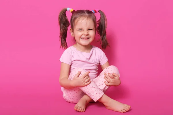 Indoor studio shot van lachen positieve Kid zittend op de vloer, poseren geïsoleerd over roze achtergrond, het dragen van Rose t shirt en broek, met ponytails, in hoge geesten. Jeugd concept. — Stockfoto