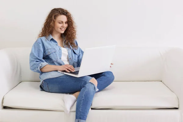 Horizontal shot of nice looking lovely attractive woman, focused curly haired girl sitting on white sofa while working remotely part time, using her modern portable computer and wireless Internet. — Stock Photo, Image