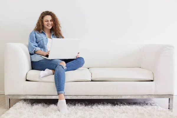 Foto de encantadora senhora satisfeita com cabelo encaracolado sentado no sofá confortável branco, usando laptop para trabalho on-line, usando Internet sem fio, estar em quarto interior leve, parece feliz e feliz . — Fotografia de Stock