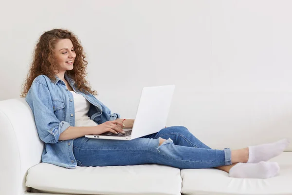 Szczęśliwy wesoły młody kobieta leżąc na jej białej kanapie, po odpoczynku, trzymając laptopa na nogach, patrząc na ekranie uważnie, wpisując informacje, surfowanie po Internecie, będąc w dobrym nastroju. Czas wolny. — Zdjęcie stockowe