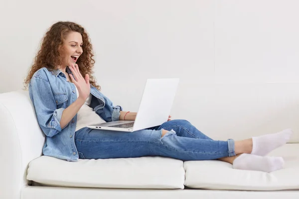 Indoor shot of emotional curly haired lady sitting on sofa, opening her mouth widely, making gesture, having videocall over laptop, time for communication in distance. People and technologies. — Stock Photo, Image
