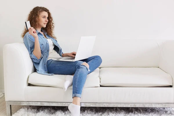 Retrato de mulher magnética pensativo sentado com seu laptop no sofá, segurando cartão de crédito em uma mão, pensando em compras on-line, como gastar dinheiro, vestindo roupas de jeans casuais. Compras . — Fotografia de Stock