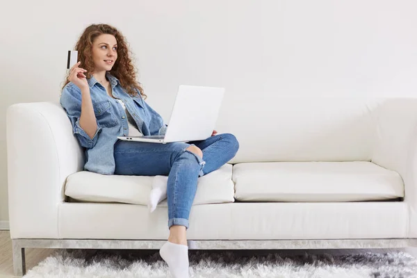 Retrato de bela senhora positiva olhando para o lado, levantando a mão com cartão de crédito, fazendo planos para compras on-line, ter laptop nas pernas, vestindo jeans e jaqueta, tendo expressão facial agradável . — Fotografia de Stock