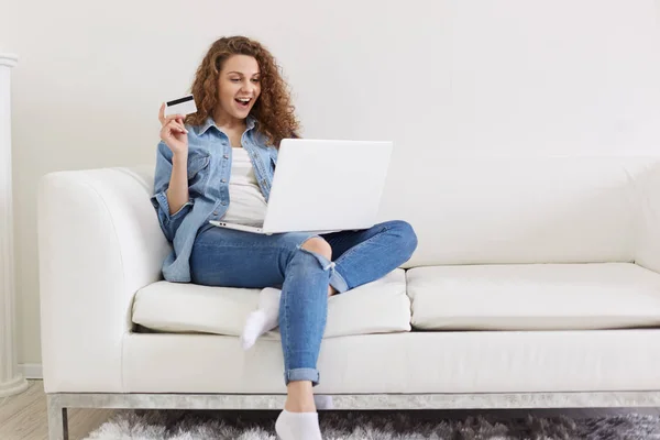 Charismatic attractive curly haired woman opening her mouth widely with satisfaction, looking at laptop screen attentively, holding credit card in hand, being concentrated on shopping. Rest concept. — Stock Photo, Image