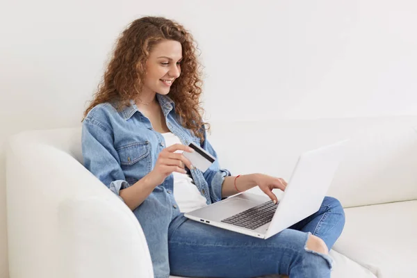 Cheerful good looking model posing on sofa at home, being busy with activities throught laptop, trying online services, smiling sincerely, holding credit card, searching for something to buy. — Stock Photo, Image