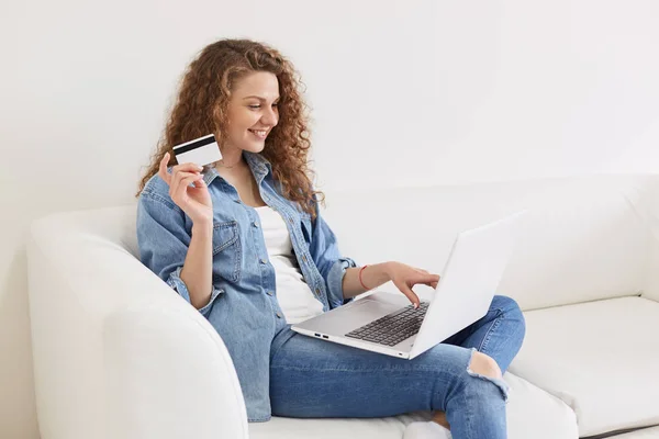 Profil wanita muda bahagia ceria dengan rambut keriting tinggal di rumah, menghabiskan hari liburnya online, akan membeli sesuatu, mengetik di laptop, memegang kartu kredit. Manusia dan teknologi konsep . — Stok Foto