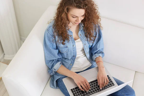 Top view of busy concentrated woman with curly hair, typing information, searching in net, looking attentively at laptop screen, being focused on her device, working online. Technologies concept. — Stock Photo, Image