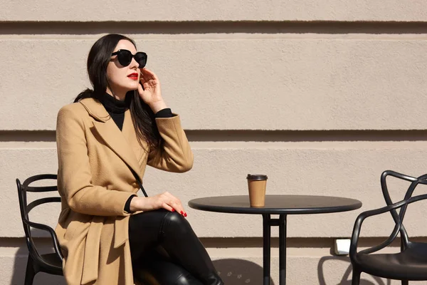 Outdoor shot of brunette woman wearing fashionable coat, black outfit and sunglasses, sitting at restaurant terrace and thinking, looking at distance, drinking takeaway coffee, having dinner break.