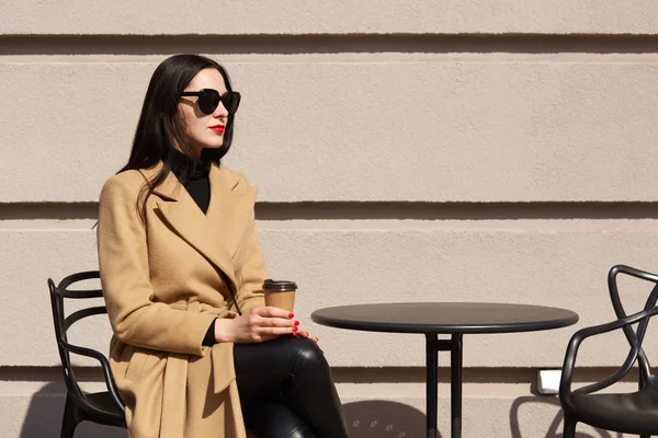 Outdoor shot of stylish woman wearing fashionable beige coat, having break in street outdoor cafe and drinking hot beverage, sitting at restaurant terrace, feels lonely, has pensive facial expression.